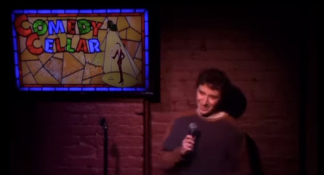 On stage with a mic at the Comedy Cellar, the stained-glass sign behind